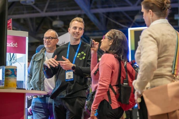 Eine Gruppe von Personen bespricht bei einer Indoor-Veranstaltung oder Messe gemeinsam Lösungen zur Flüssigkeitszufuhr. Im Hintergrund sind Ausstellungsstände und Produkte zu sehen.